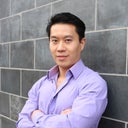 Portrait of Dr. Tsai. He is wearing a lavendar button up shirt and leaning against a brick wall with arms folded.