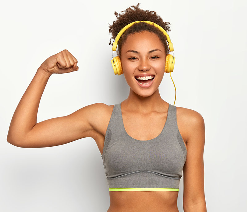 Horizontal view of dark skinned woman in good mood, raises arm with muscles, has strong body, dressed in gym outfit, listens audio via modern headphones, poses indoor. Fitness and music concept